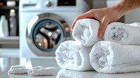 Fresh white rolled towels stacked by washing machine, with hand arranging clean laundry, showing modern home lifestyle and housekeeping routine.