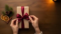 Hands tying burgundy ribbon bow on cream gift box with pine branches and dried orange slices on wooden table for holiday wrapping celebration.