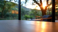 Wooden table surface at sunset with blurred nature background through window, golden sunlight creating warm atmosphere on empty tabletop, peaceful evening mood.