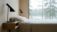 Serene bedroom interior with minimalist design, modern black task lamp on wooden nightstand, white bedding, and forest view through large window.