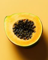 Fresh cut papaya half showing bright orange flesh and black seeds against yellow background, close-up view for food and healthy eating concepts.