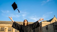 Young Asian woman graduate celebrating with arms raised throwing mortarboard cap in air against blue sky and university building background.