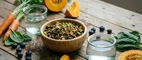 Wooden bowl filled with mixed seeds and grains surrounded by fresh vegetables, citrus fruits, water glasses, and blueberries on rustic wooden surface for healthy eating and nutrition content.