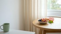 Wooden round table with bowl of granola, fresh grapefruit slice, mint leaves, and green mug by window with beige curtains for breakfast and lifestyle content.