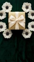 Golden gift box with cream ribbon surrounded by white anemone flowers on dark green velvet background for holidays and special occasions.