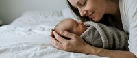 Tender moment of loving mother holding sleeping newborn baby wrapped in soft knit blanket on white bed. Maternal bonding and early parenthood concept.