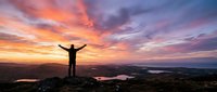 Person with raised arms celebrating freedom and success on mountain peak during dramatic sunset with colorful sky over landscape with lakes below.
