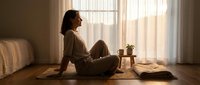 Young woman in cozy sweater sitting on wooden floor by window with warm sunlight, peaceful morning meditation and relaxation at home.