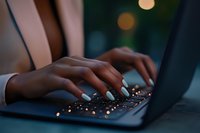 Female hands with blue manicure typing on illuminated laptop keyboard in dark room with bokeh lights background, creating atmospheric night work environment.