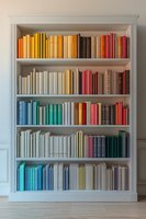 Books arranged by color spectrum on white wooden bookshelf creating rainbow pattern. Classic hardcover volumes organized aesthetically on library shelves against wall.