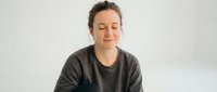 Peaceful young woman with closed eyes meditating in comfortable gray sweater against clean white background for wellness and mindfulness content.