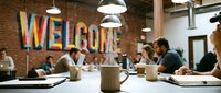Steaming coffee mug on a table in a modern coworking space with a colorful welcome sign on a brick wall and people working.