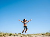 Young child with curly dark hair and medium skin tone jumping with arms raised in celebration on sandy beach terrain under clear blue sky, expressing joy and freedom outdoors.