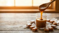 Homemade salted caramel sauce dripping from wooden spoon into glass jar surrounded by soft caramel candies on rustic wooden table with warm sunlight.
