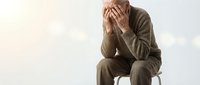 Stressed businessman sitting on chair covering face with hands showing depression, anxiety and mental health struggles in professional workplace environment.