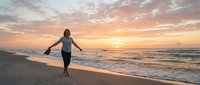 Woman in wetsuit running into ocean waves at golden hour sunrise, capturing active beach lifestyle and water sports enthusiasm.