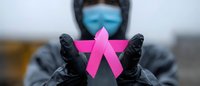 Healthcare worker in protective mask and black gloves holds pink breast cancer awareness ribbon against blurred background, symbolizing support and medical care.