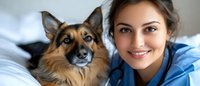 Young female veterinarian in blue scrubs smiling with German Shepherd dog patient in clinic examination room for pet healthcare and animal medicine.
