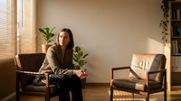 Young Asian woman sitting relaxed in modern living room with houseplants and natural sunlight streaming through window blinds creating warm atmosphere.