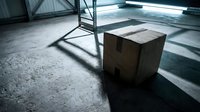 Cardboard box on concrete warehouse floor with dramatic shadows from industrial shelving and natural light streaming through windows.