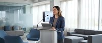 Professional businesswoman presenting at podium in modern office conference room with laptop and microphone for corporate meetings.