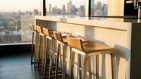 Modern bar counter with wooden stools in high-rise office overlooking city skyline at golden hour, contemporary workspace design.