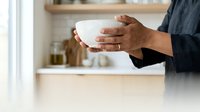 Person of South Asian descent holding empty white bowl in modern kitchen, wearing dark shirt. Suitable for food service, cooking, recipes, meal preparation, and culinary content.