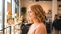 Young Caucasian woman with wavy blonde hair in coral sweater smiling in modern cafe or restaurant interior with warm lighting and plants.