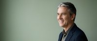 Mature man with gray hair and light skin tone wearing dark blazer over casual shirt, smiling at camera against neutral green background for corporate portraits and business headshots.