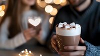 Hot chocolate topped with marshmallows in glass mug held by person wearing sweater, romantic bokeh lights and heart shape in background, cozy winter mood.