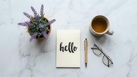 Flat lay of a notebook with hello lettering, lavender plant, tea cup, pen and glasses on marble surface.