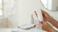 Close-up of hands holding white cosmetic jar in bright bathroom setting with skincare products blurred in background for beauty and wellness content.