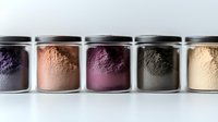 Glass jars filled with colorful superfood powders in purple, pink, burgundy, green and beige shades lined up against white background for healthy eating concept.