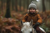 Young child wearing winter clothing and knit hat riding white horse through autumn forest, capturing innocent joy and outdoor adventure in natural setting.