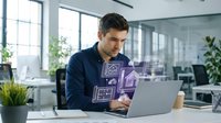 Young Caucasian businessman working on laptop with digital holographic interface displaying smart home automation icons in modern office environment.
