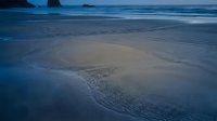 Serene ocean beach at twilight with gentle waves creating rippled sand patterns and distant rock formations silhouetted against moody blue sky for travel content.