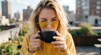 Young blonde woman in yellow sunglasses drinking coffee from black cup on urban rooftop terrace with city skyline background during golden hour lifestyle moment.