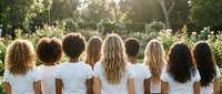 Diverse group of women with different hair textures and ethnicities standing together in white shirts outdoors during golden hour sunlight.