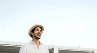 Young adult male with dark hair and Mediterranean ethnicity wearing a light beige straw hat and white linen shirt, smiling outdoors against modern architecture backdrop.