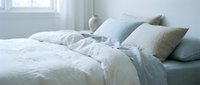 Unmade bed with white and blue linens, decorative pillows, and natural light from window, suitable for home decor and lifestyle content.