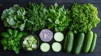 Fresh green vegetables and herbs arranged in row on dark wooden background. Lettuce, dill, basil, cucumber slices and purple onion dip for healthy eating.