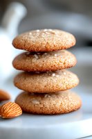 Stack of fresh baked almond cookies with sea salt sprinkled on top, macro shot showing texture detail on white surface with natural lighting.