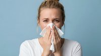 Young adult woman with fair skin and blonde hair pulled back, wearing a white shirt, holding a tissue to her nose against a blue background, depicting cold or allergy symptoms for healthcare and wellness content.