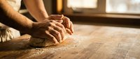 Male baker hands kneading fresh dough on wooden surface in warm kitchen light for homemade bread making and artisan baking preparation.