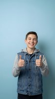 Happy teenage boy showing thumbs up gesture in denim jacket against blue background expressing positive emotions and success for youth lifestyle concepts.