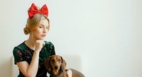 Young woman with blonde hair and fair complexion wearing red bow headpiece and dark sequined top, posing thoughtfully with brown dachshund dog against white background for holiday or festive themed designs.