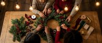 Family creating festive Christmas wreath together at wooden table with pine branches, berries, pinecones and warm string lights overhead during holiday crafting.