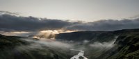 Dramatic mountain valley landscape with winding river, misty fog, and golden sunlight breaking through storm clouds at dawn or dusk for nature and travel projects.