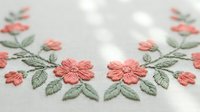 Delicate coral pink floral embroidery with green leaves on white fabric. Hand-stitched decorative border pattern for textile crafts and sewing projects.