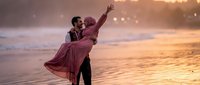 Romantic couple dancing on beach at sunset with golden light and ocean waves, man lifting woman in flowing dress for wedding or engagement celebration.